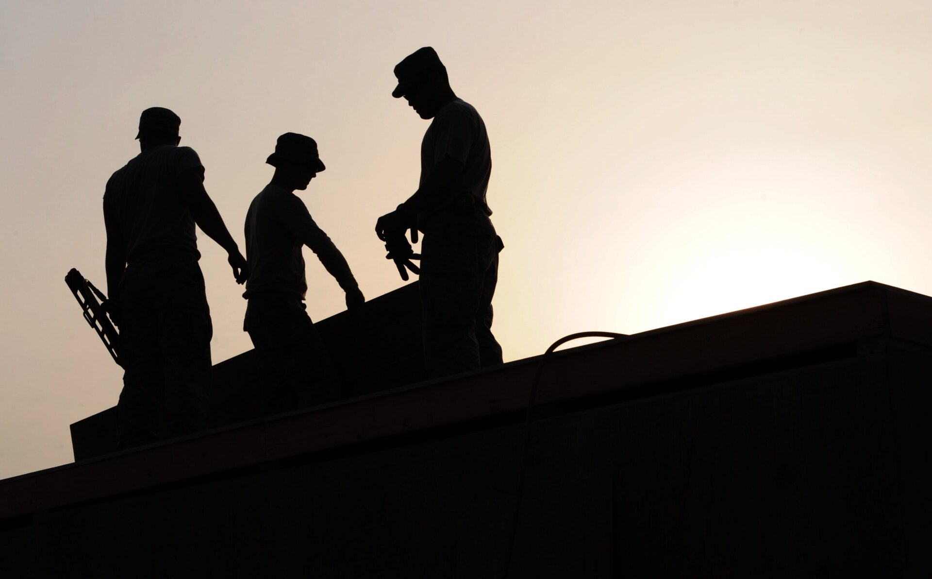 Silhouettes of three men working on top of a roof as the sun sets behind them.